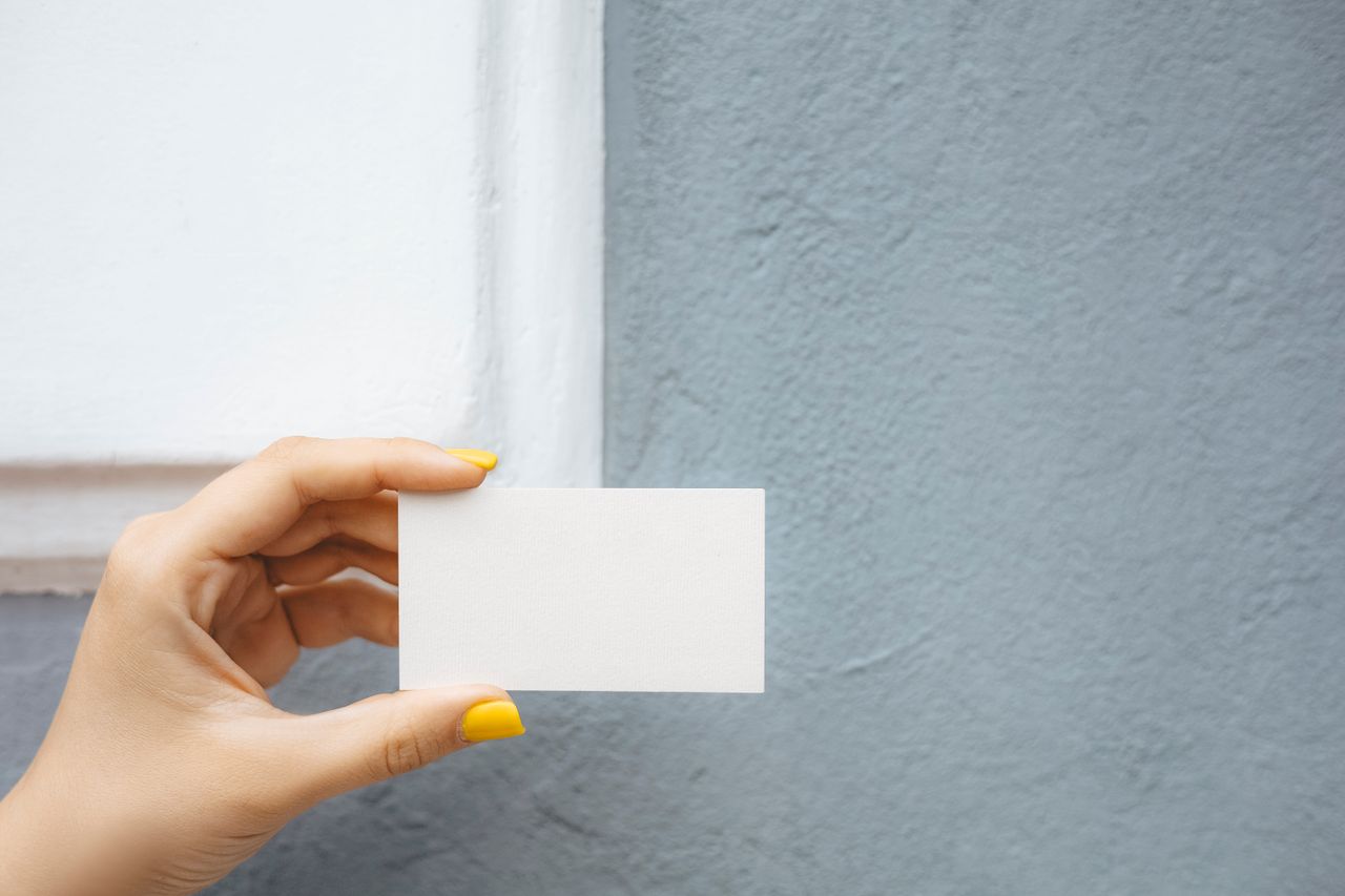 Close-up of a hand with yellow nail polish holding a blank white card against a textured blue wall.